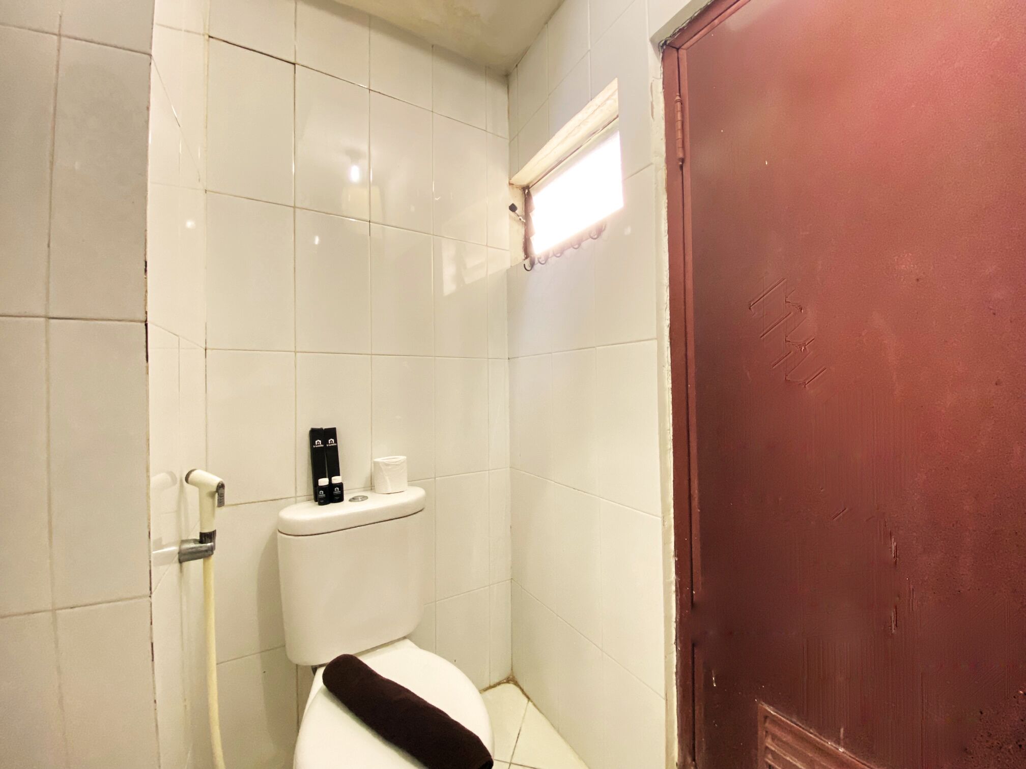 A small, compact bathroom with white tiles, a white toilet, a black towel on the seat, and a red door.