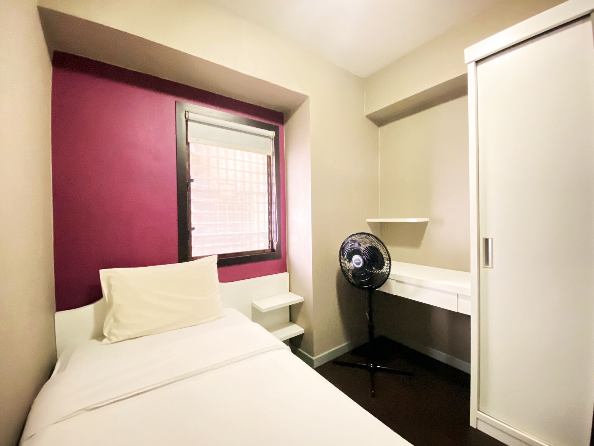 Una habitación de hotel con cama, ventana con persianas, un ventilador de pie y un armario blanco.