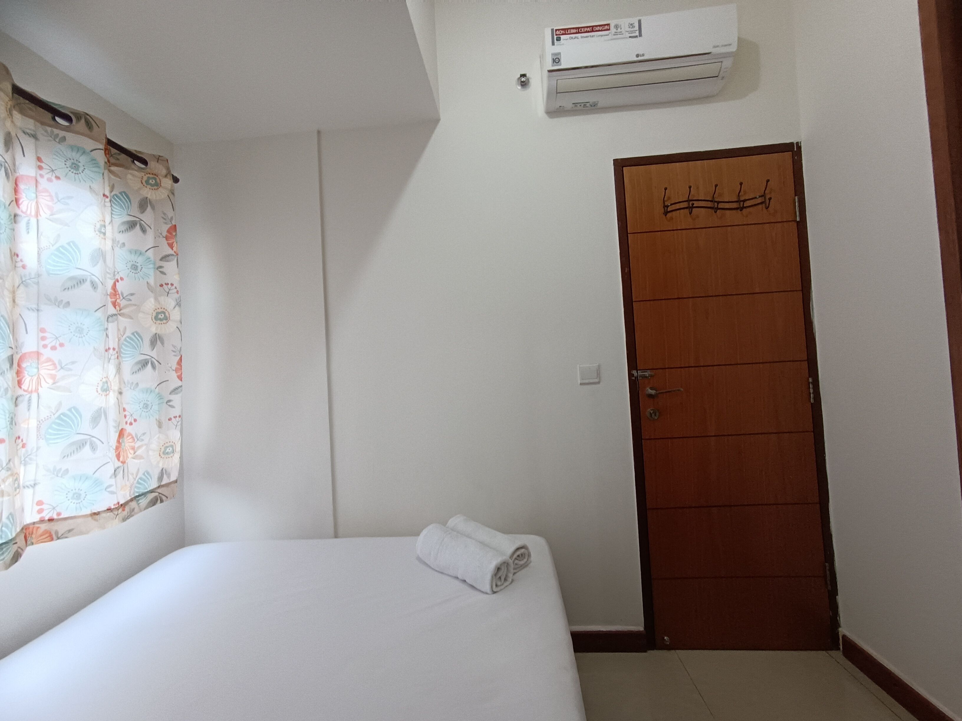 A small, single-bed room with a wooden door, a window with floral curtains, and an air conditioning unit on the wall.