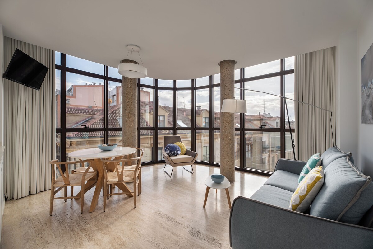 A modern living room with a round dining table, a grey sofa, and a large window offering a city view.