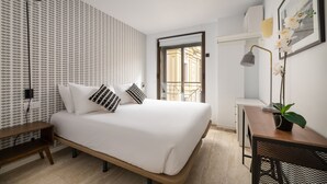 Un dormitorio moderno con una cama grande, un escritorio y una silla. Se ve una ventana con vista a un edificio.