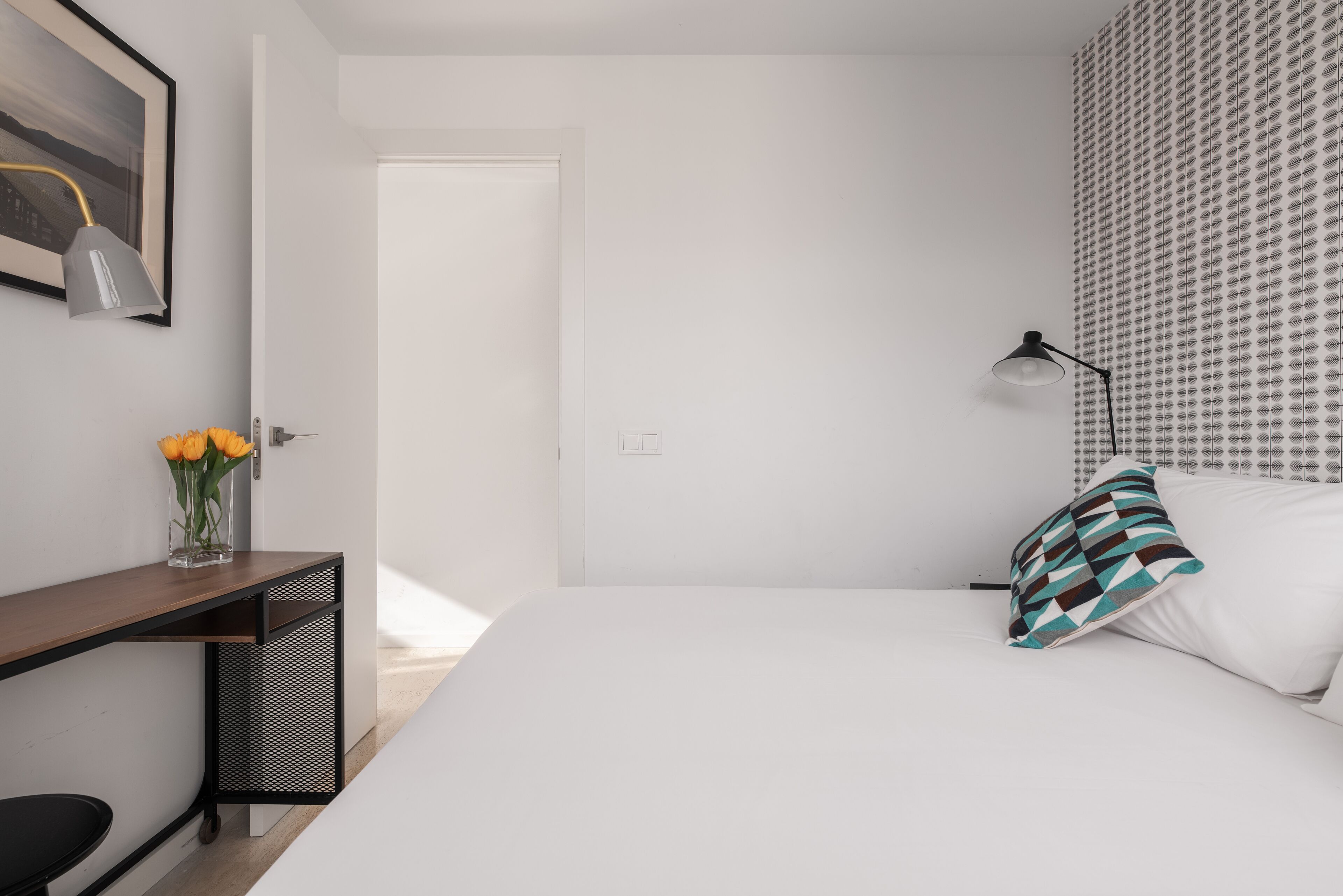 Un dormitorio moderno con una cama, un escritorio, una silla, una lámpara y un papel tapiz estampado.