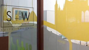 Una ventana de vidrio con el texto "S+W A STORGA SUITE THE WAY" y el reflejo de una estructura amarilla con el perfil de un edificio.