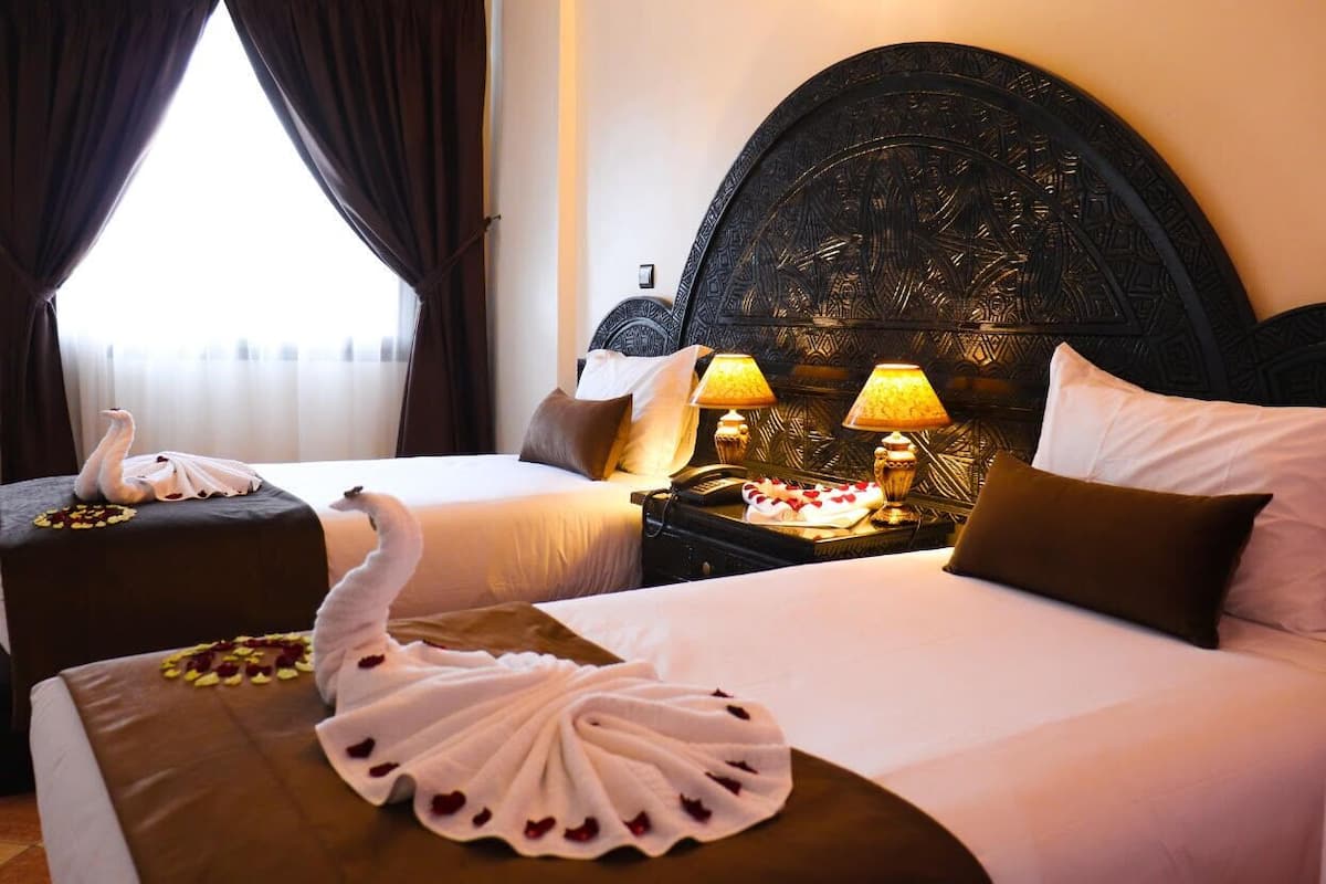 A hotel room with two beds, a decorative headboard, bedside lamps, and a swan-shaped towel on the beds.