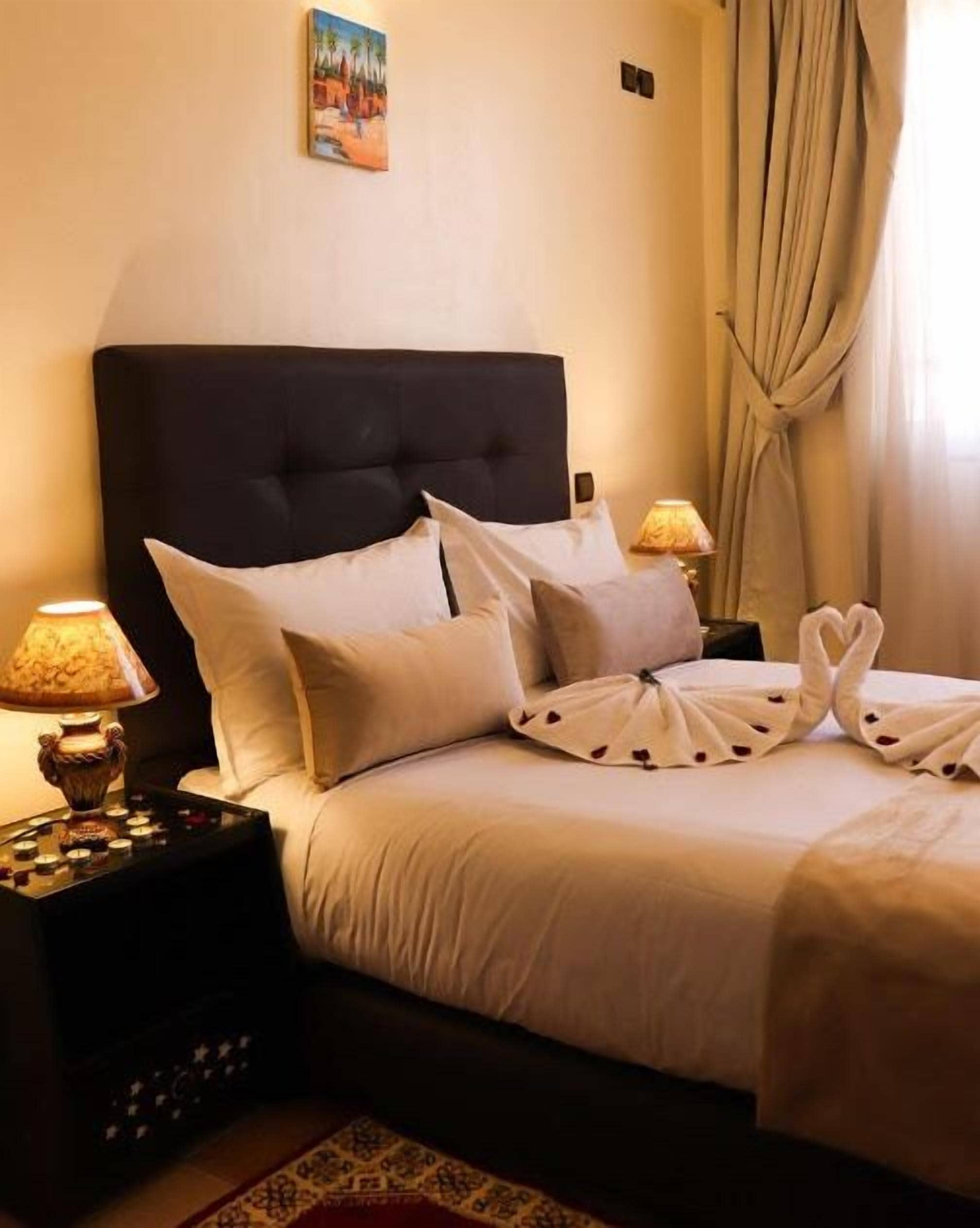 A neatly made bed with white linens and decorative pillows, a bedside table with a lamp, and a painting on the wall.