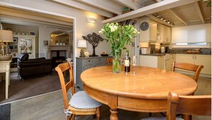 Un coin repas avec une table en bois, des chaises et un vase de fleurs. On aperçoit, au fond, une cuisine avec des placards et une horloge.