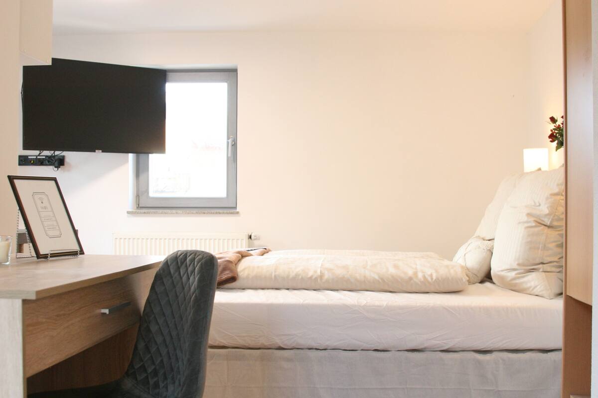 A bedroom with a bed, a desk, a chair, a television mounted on the wall, and a window.