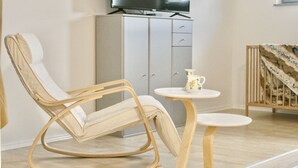 A room with a beige rocking chair, a small round table with a white pitcher, and a grey cabinet with a television above it. - The Manhattan A Boutique Hotel By American Ease (Grafenwohr)