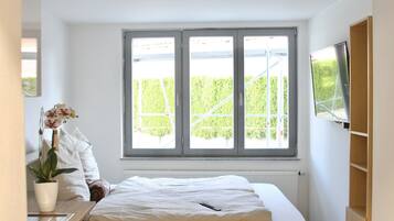A bedroom with a large window, a bed, a bedside table with a plant, and a TV mounted on the wall.