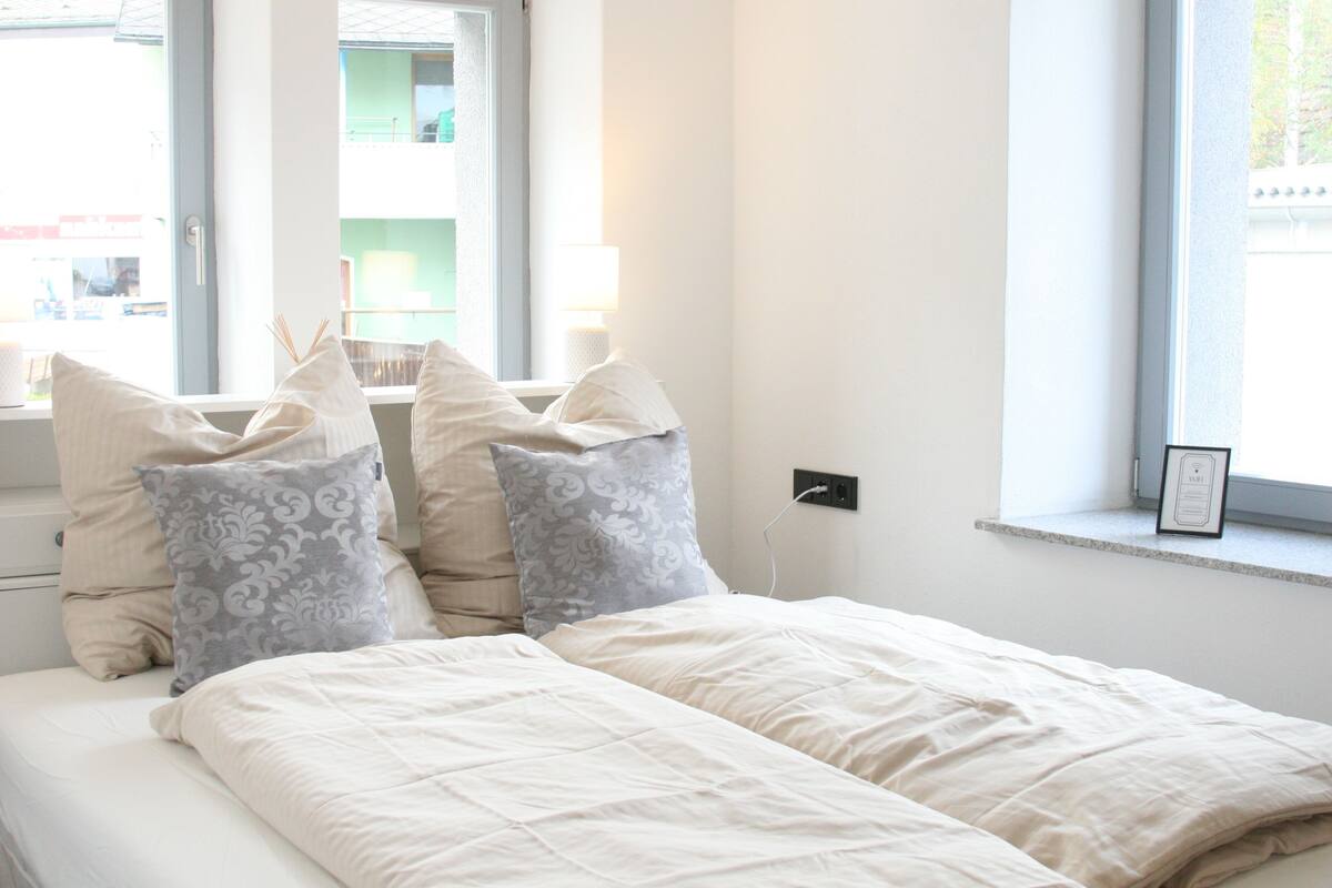A bedroom with a bed, pillows, a window, and a wall socket.