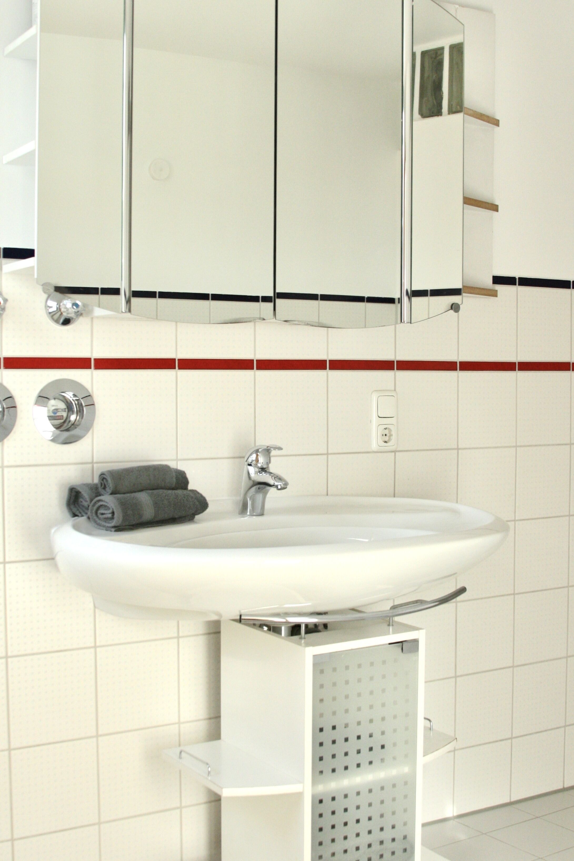 A bathroom with a white sink, chrome faucet, and a mirror above it. There is a towel rack with towels and a small shelf below the sink.