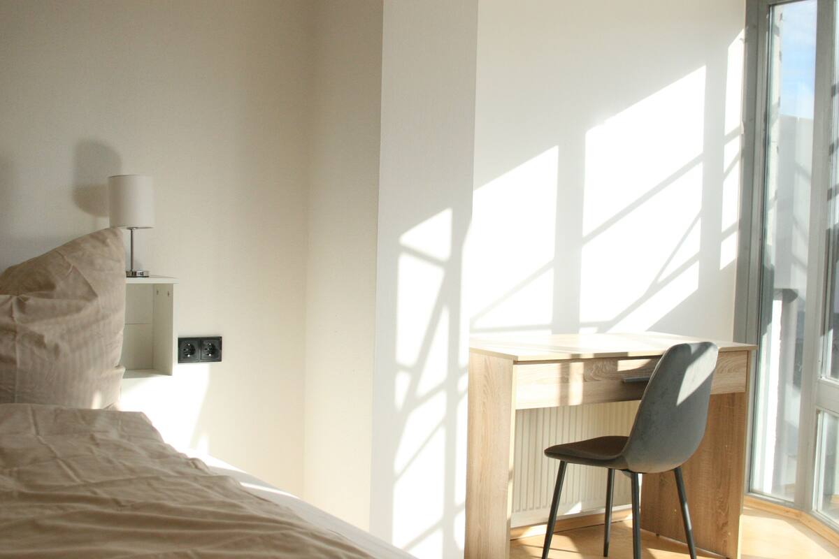 A bedroom with a bed, a desk, and a chair. There is a window providing natural light and a view outside.