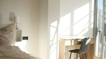 A bedroom with a bed, a desk, and a chair. There is a window providing natural light and a view outside.