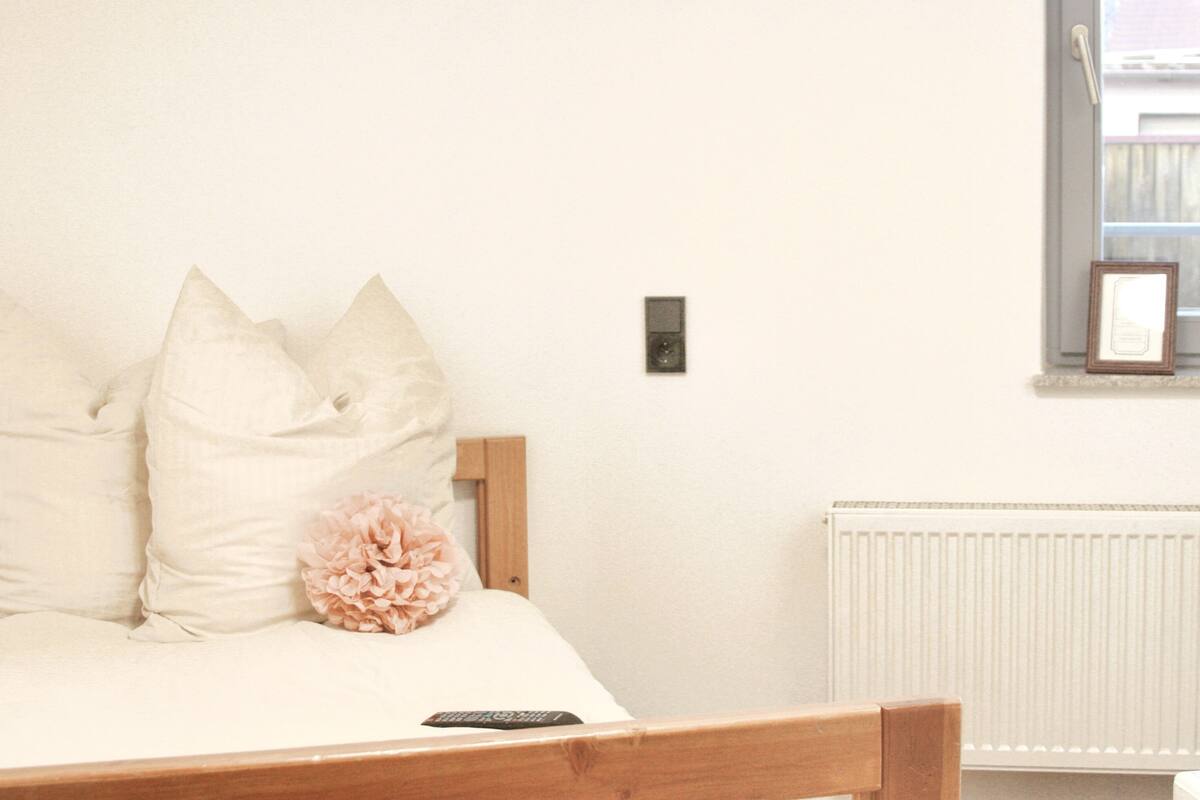A bedroom with a bed, pillows, a remote control, a wall-mounted light switch, and a radiator.