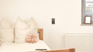 A bedroom with a bed, pillows, a remote control, a wall-mounted light switch, and a radiator.
