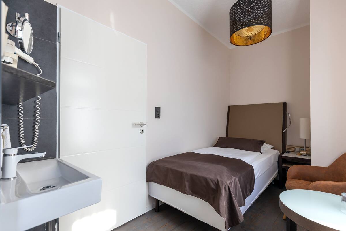 A hotel room with a bed, a bedside table, a chair, a desk, a lamp, a mirror, and a towel rack.