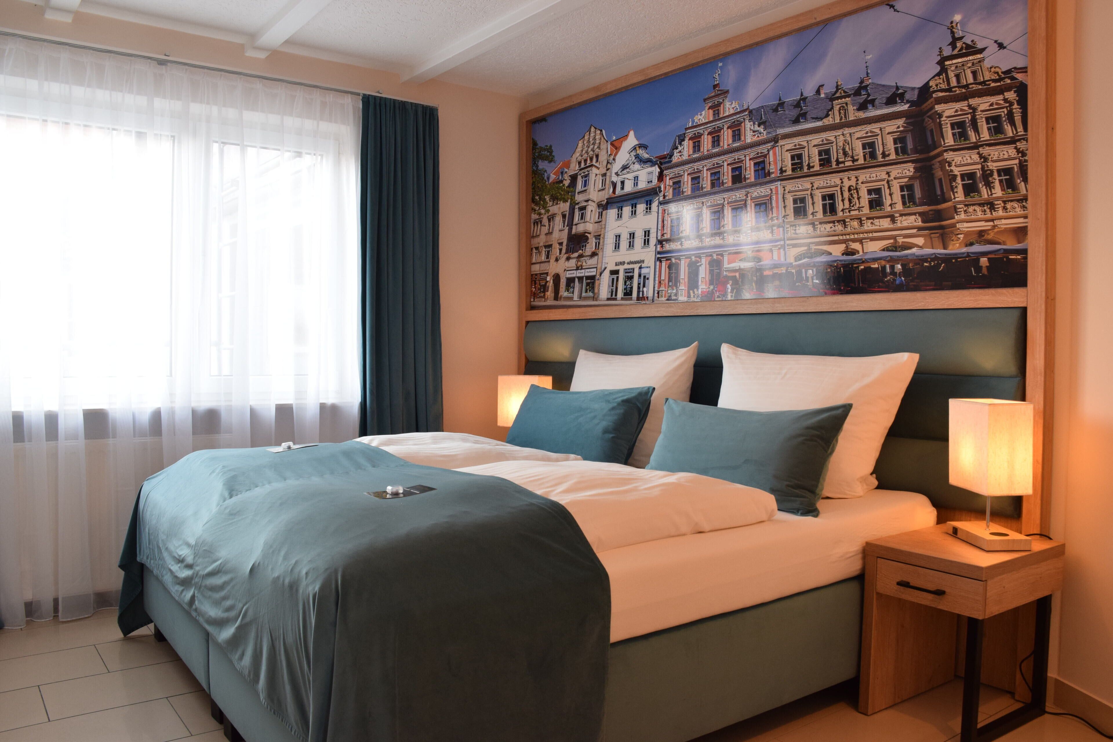 Una habitación de hotel con una cama grande, cabecera con un mural de paisaje urbano, dos lámparas de noche y una ventana con cortinas translúcidas.