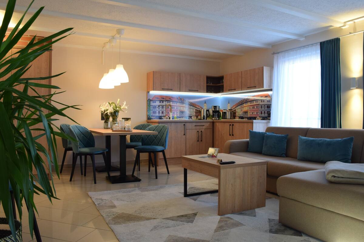 A modern living room with a sofa, coffee table, and dining area. There is a kitchen in the background with wooden cabinets and a view of buildings.