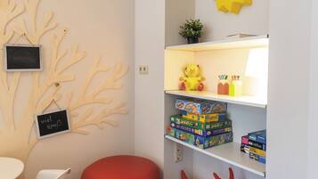 Ein Zimmer mit weißer Wand, einer Holz-Baumdekoration, einem roten Pouf, einem Regal mit Spielzeug und Spielen sowie einer kleinen Tafel mit Text.