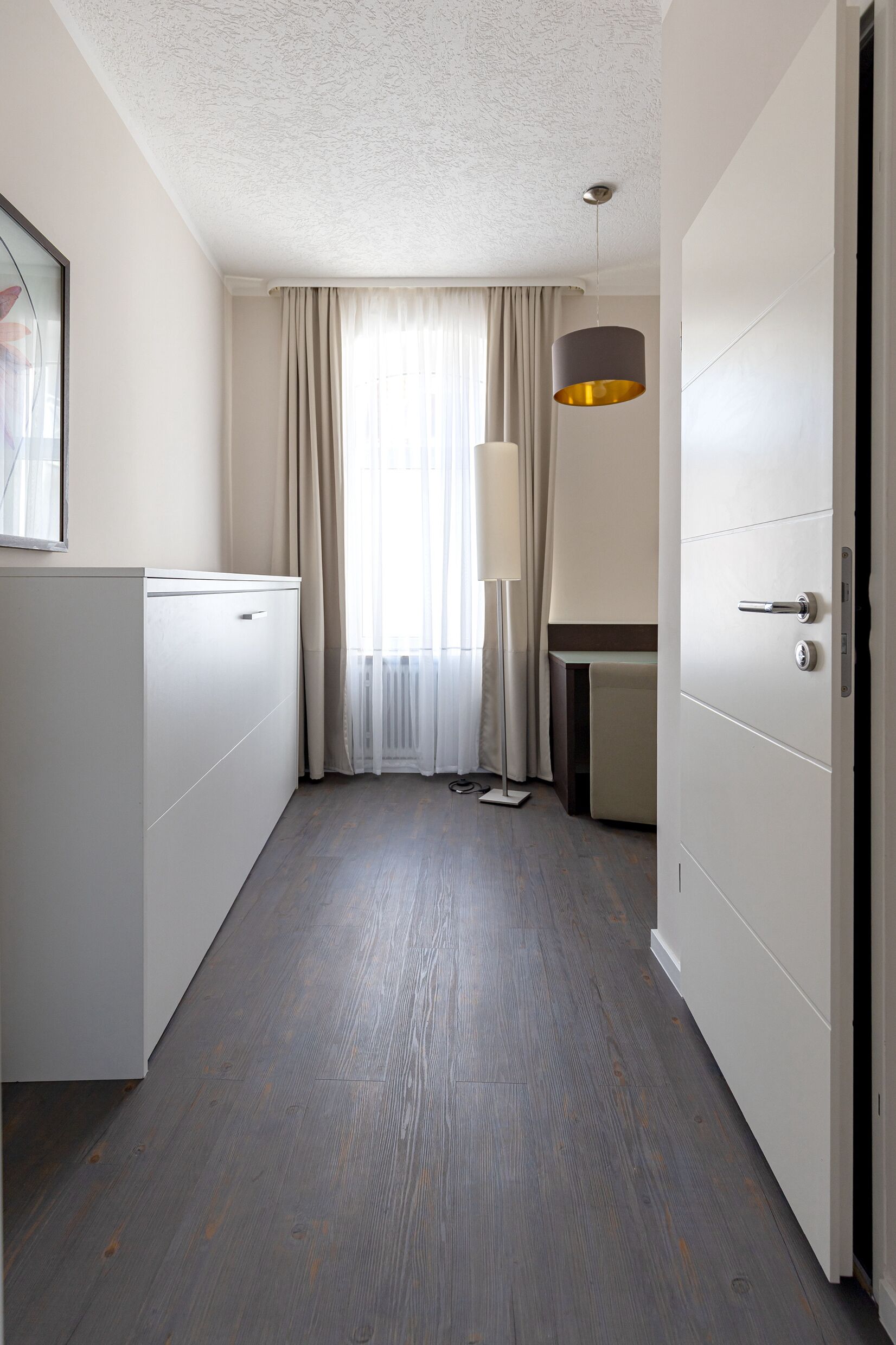 Una habitación de hotel con piso de madera, una cama, un armario, un escritorio y una ventana con cortinas.