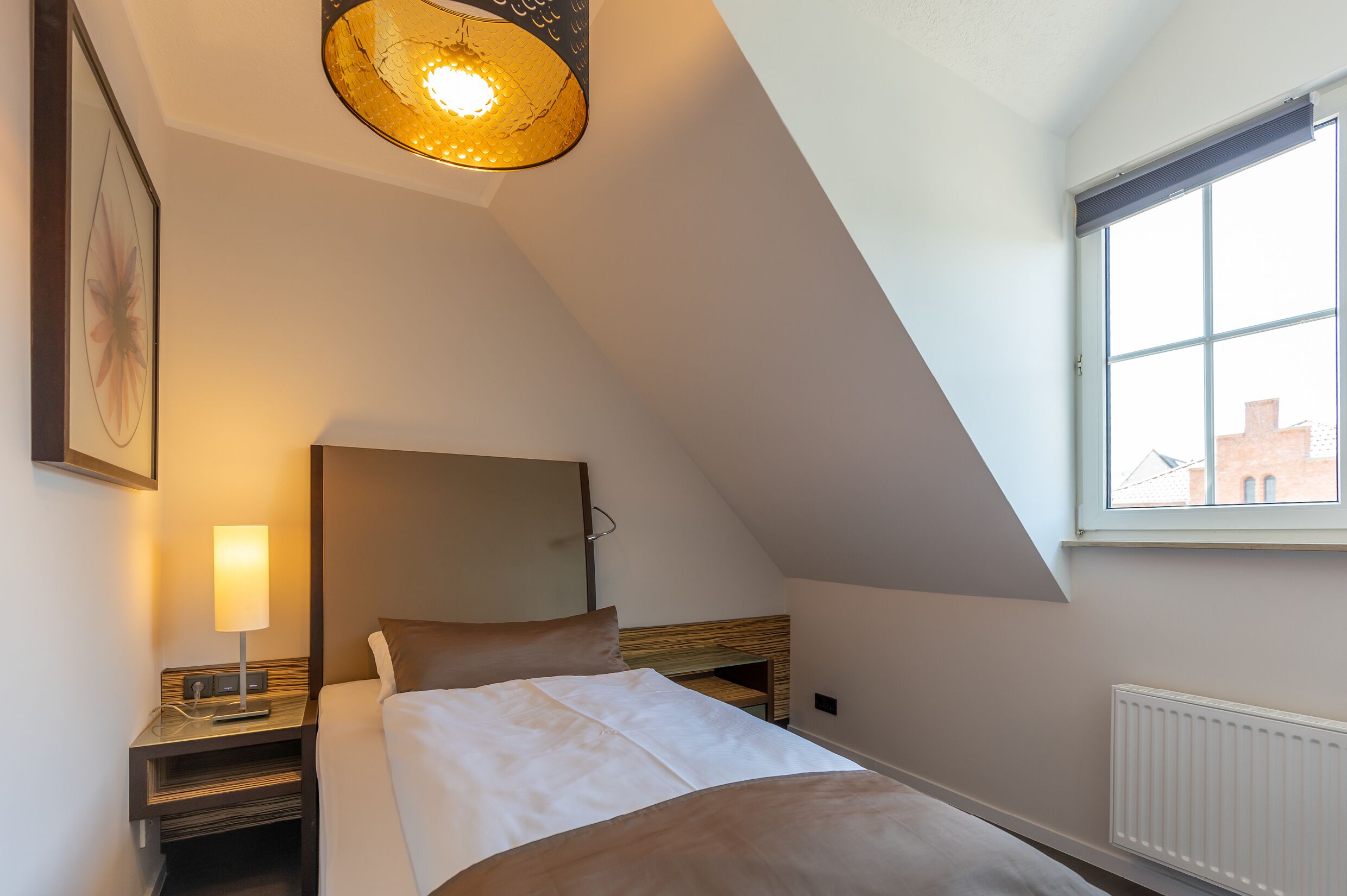 A bedroom with a bed, bedside table, lamp, and a large window with a view of buildings.