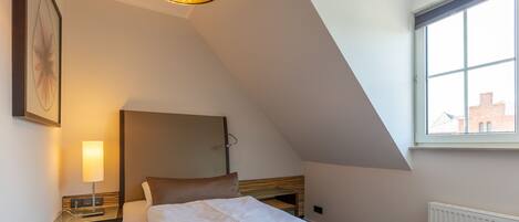 A bedroom with a bed, bedside table, lamp, and a large window with a view of buildings.