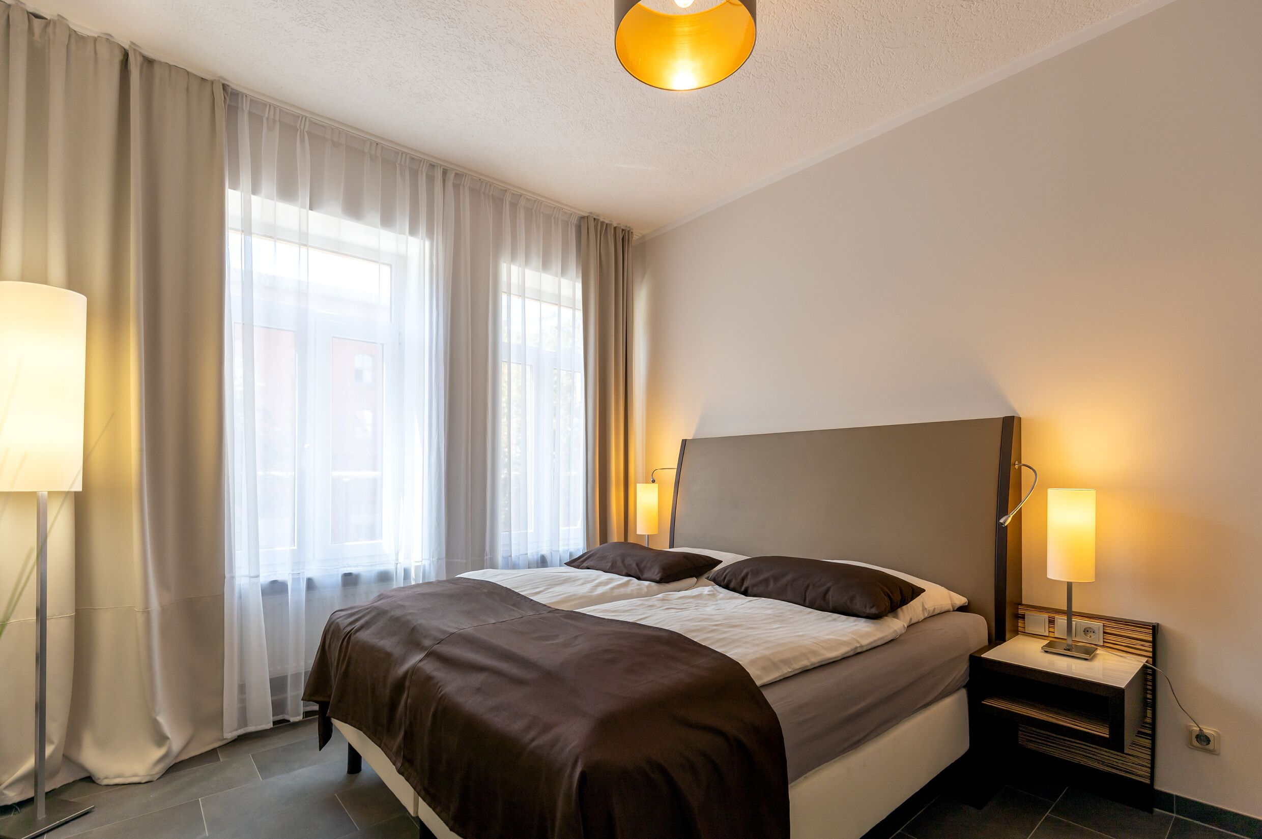 Ein Hotelzimmer mit einem Bett, zwei Nachttischlampen, einem Nachttisch und einem Fenster mit Vorhängen.