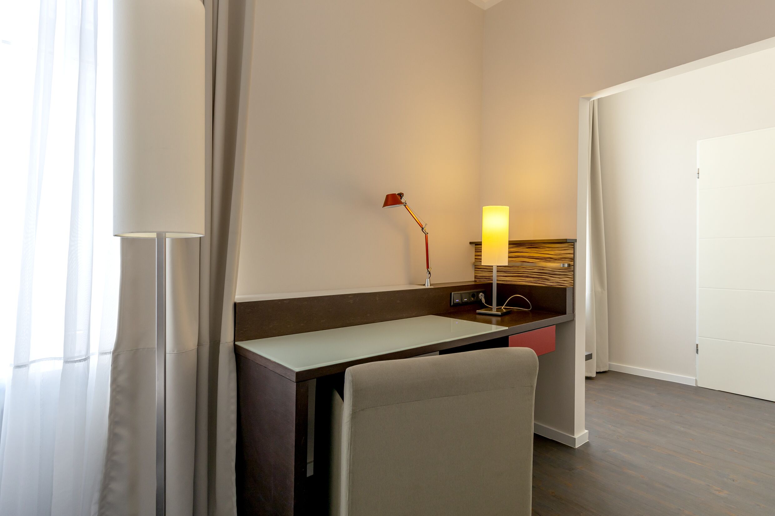 A hotel room with a desk, chair, lamp, and a door.