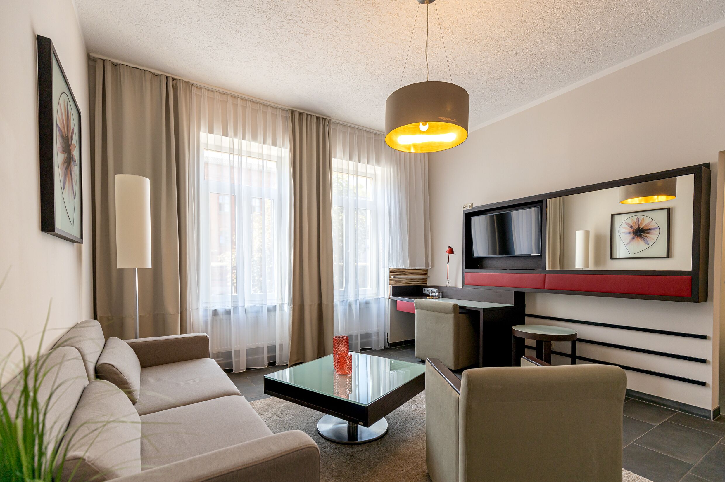 A modern hotel room with a sofa, a glass coffee table, a TV, and a dining area.