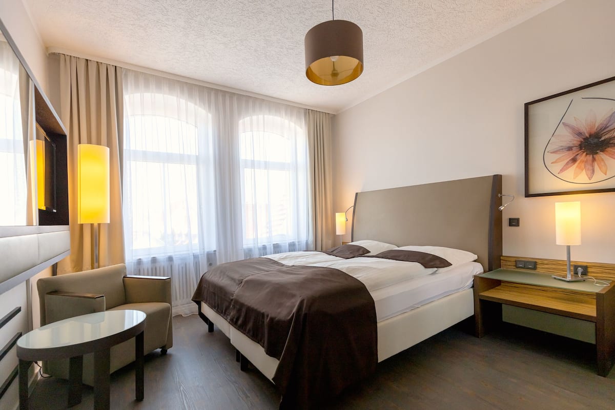 A hotel room with a bed, bedside tables, a chair, a desk, and a large window with curtains.