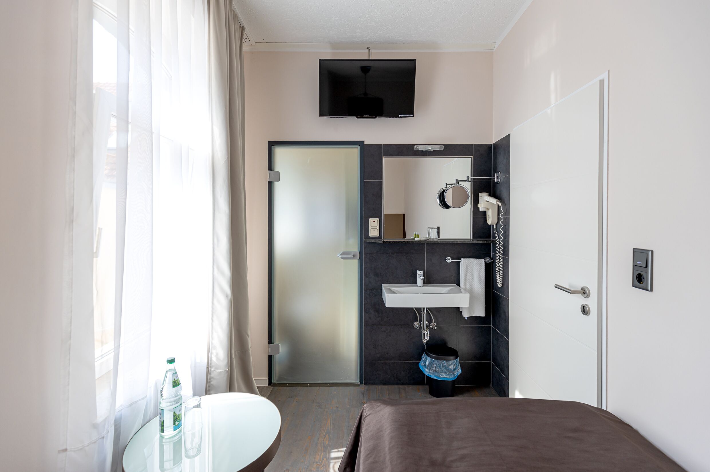 Habitación de hotel moderna con cama, una mesa redonda, una botella, un lavamanos blanco y zona de ducha.