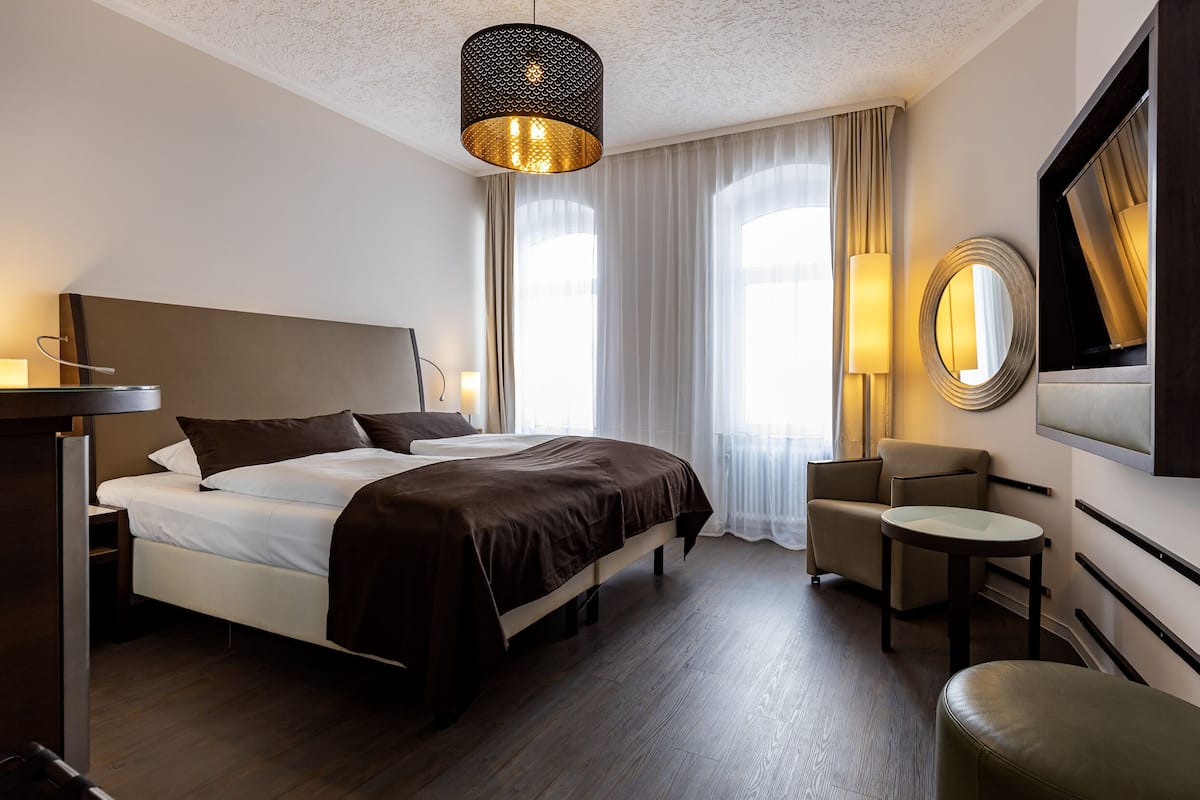 A hotel room with a large bed, a bedside table, a chair, a round mirror, and a hanging light fixture.