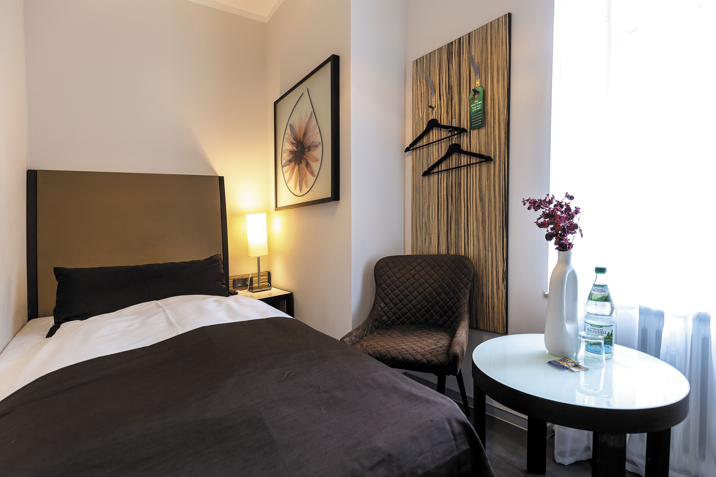 A hotel room with a bed, a chair, a small table with a vase and water bottle, a wall-mounted coat rack, and a framed picture.
