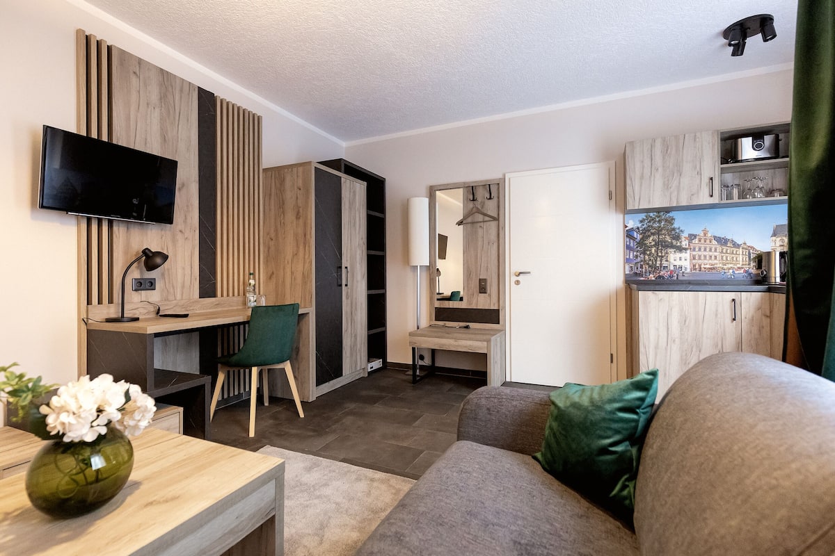 A modern living room with a flat-screen TV, wooden panel walls, a desk with a chair, a sofa, and a mirror reflecting a cityscape.
