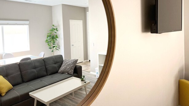A modern living room with a grey sofa, a white coffee table, and a large mirror reflecting the room. - Room in House - Cheerful And Cozy one Large Bedroom (Jurupa Valley)