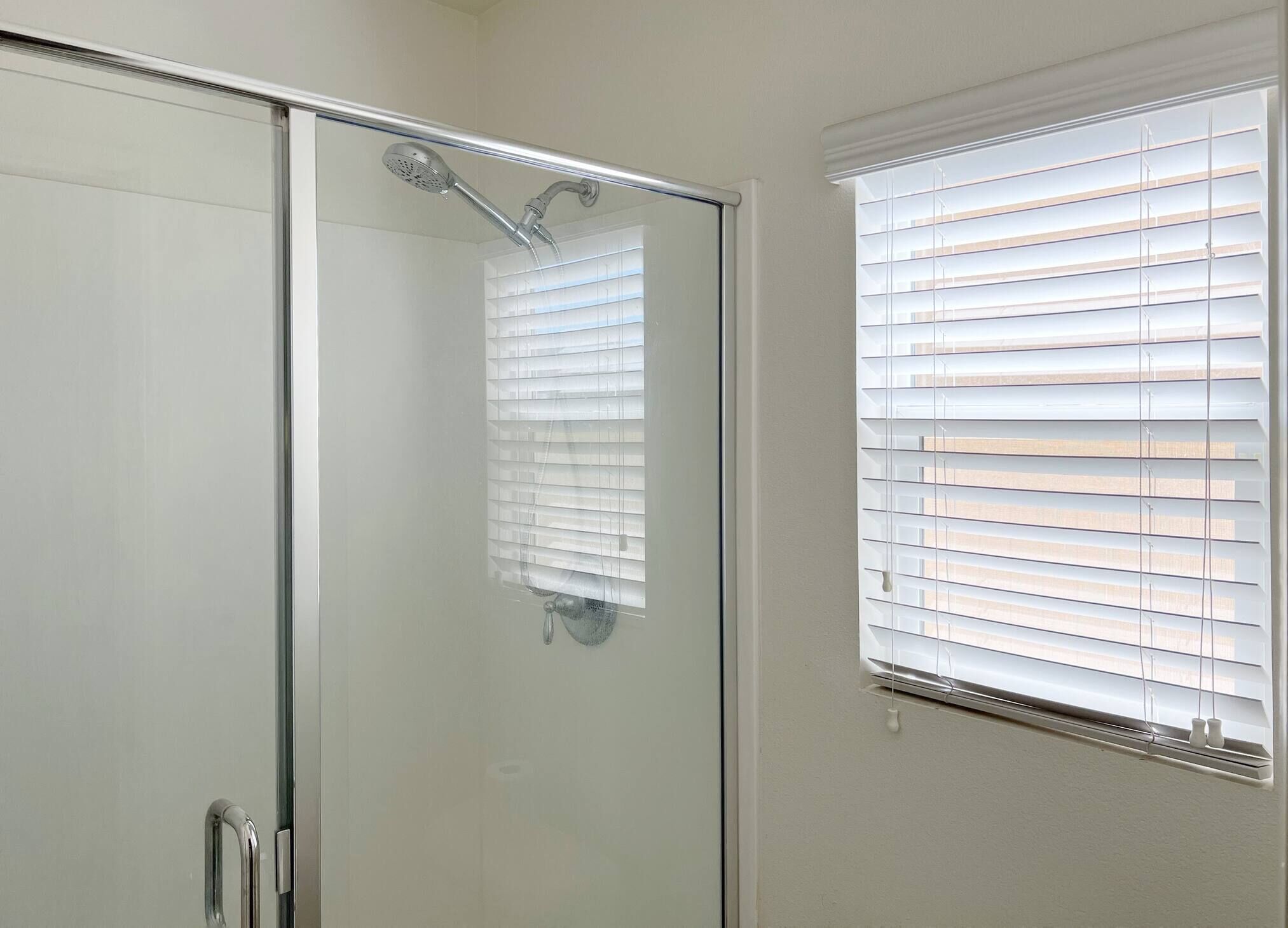 A glass-enclosed shower with a handheld showerhead, a glass door, and a window with blinds.