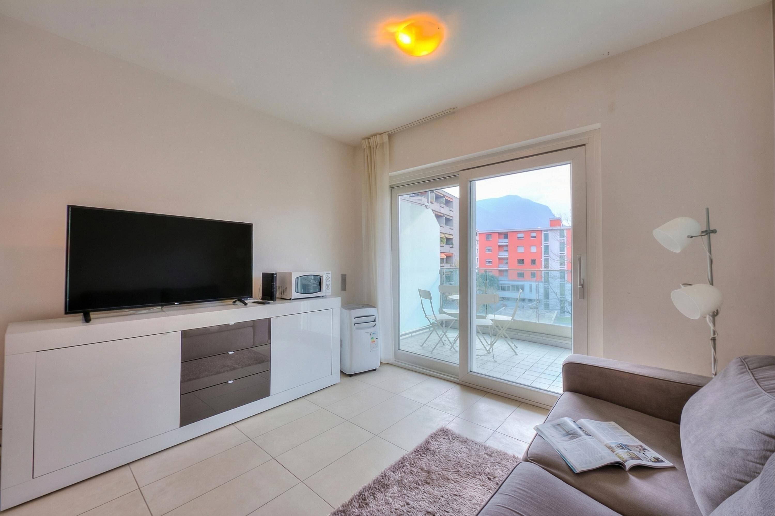 A modern living room with a flat-screen TV, a microwave, a small refrigerator, a sofa, and a large sliding glass door leading to an outdoor balcony.