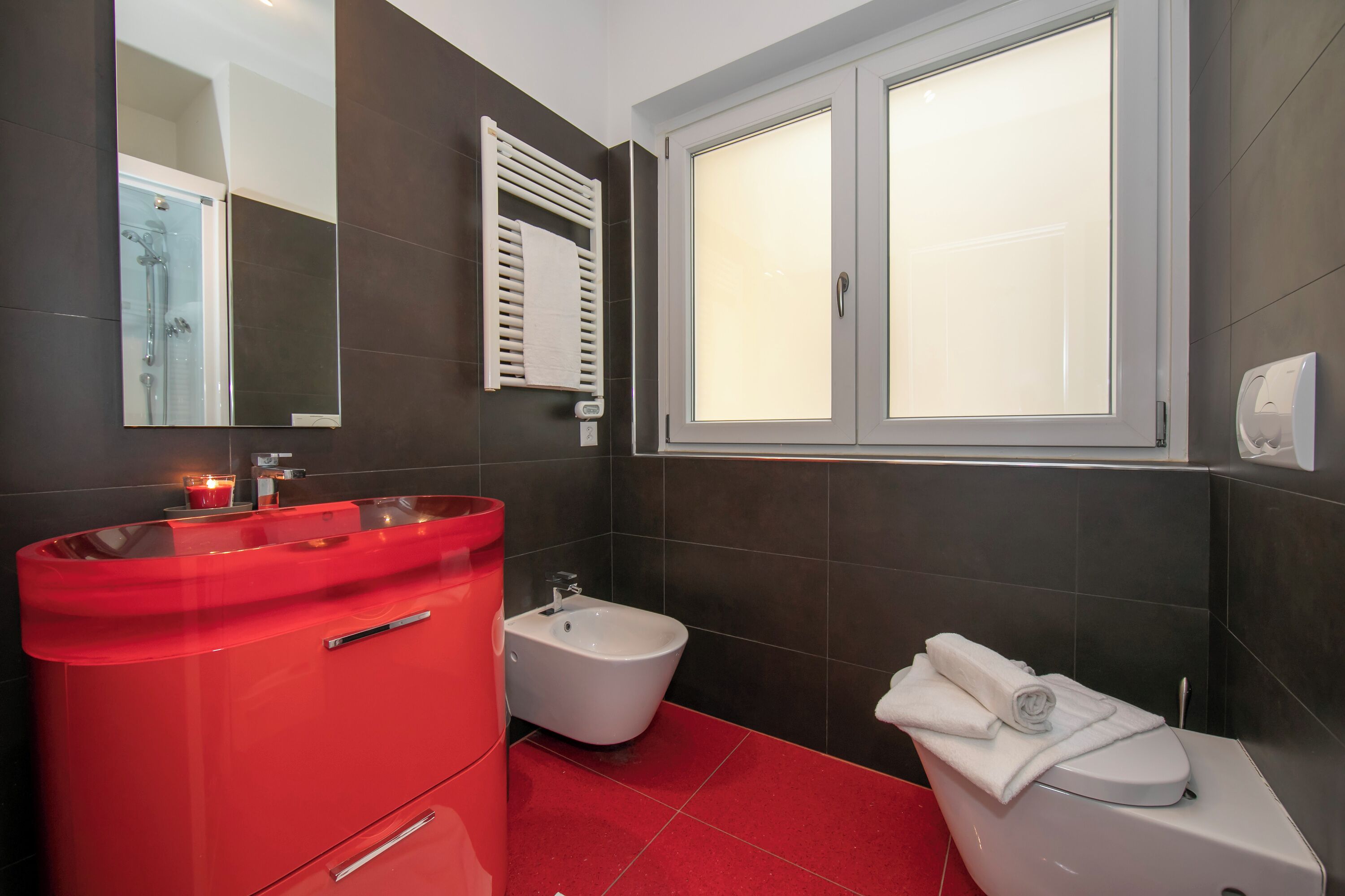 Un baño con tocador rojo, un bidé, un inodoro y una ventana con vidrio esmerilado.