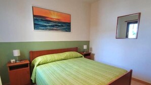 Une chambre à coucher comprenant un lit, une table de chevet, un miroir fixé au mur et un tableau représentant un coucher de soleil sur l’océan.