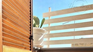 Un petit balcon fermé, doté d’une porte à persiennes en bois, d’un pot blanc posé sur le rebord et offrant une vue sur une clôture en bois et des cactus.