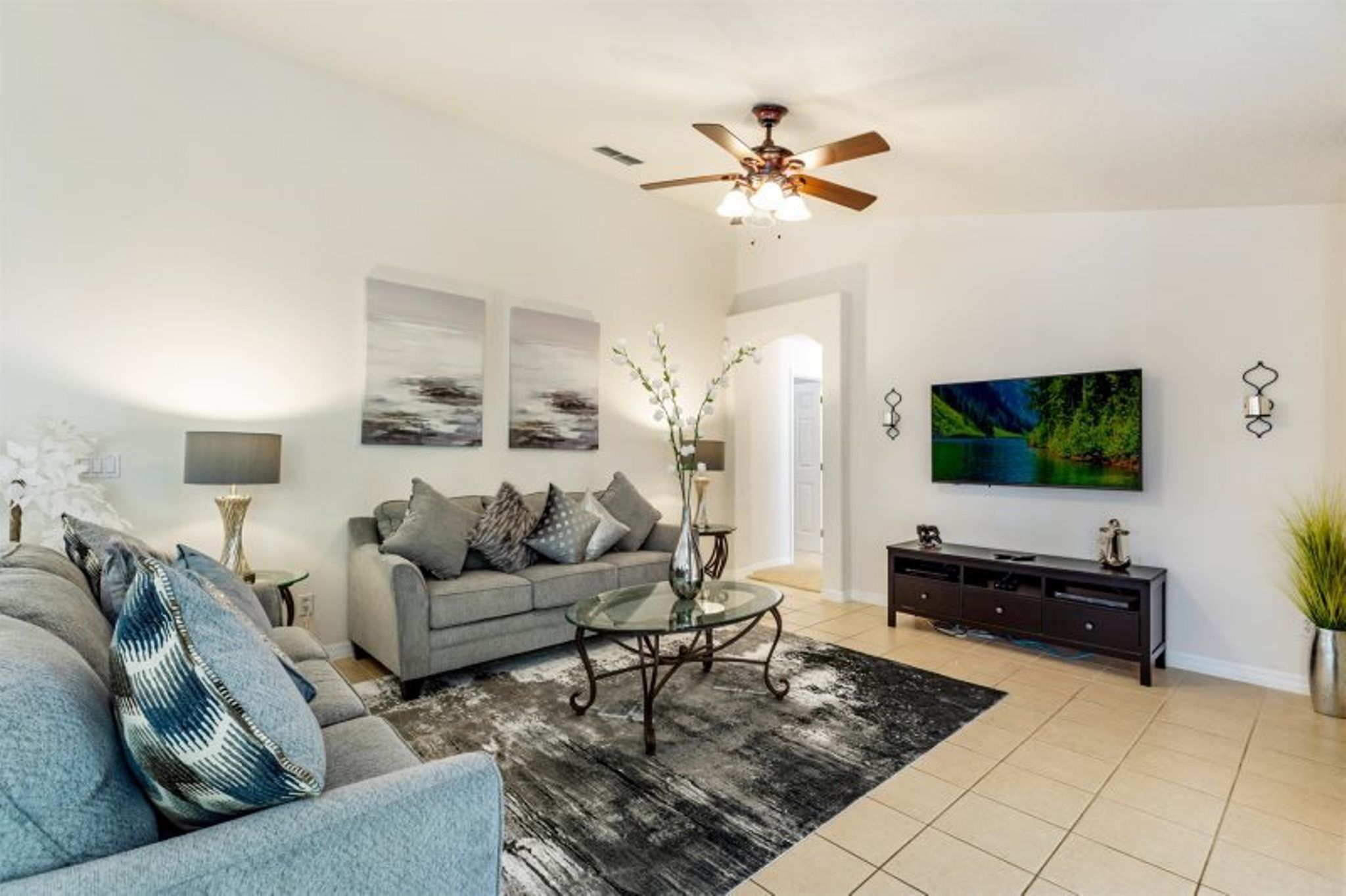 A living room with a sofa, coffee table, and TV. There is a ceiling fan, artwork on the walls, and a plant.