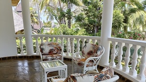 A covered outdoor seating area with white wicker furniture and a glass table, surrounded by a white balustrade and lush tropical vegetation. - 5-bedroom Villa - 3 Mins Walk To The Beach (Diani Beach)