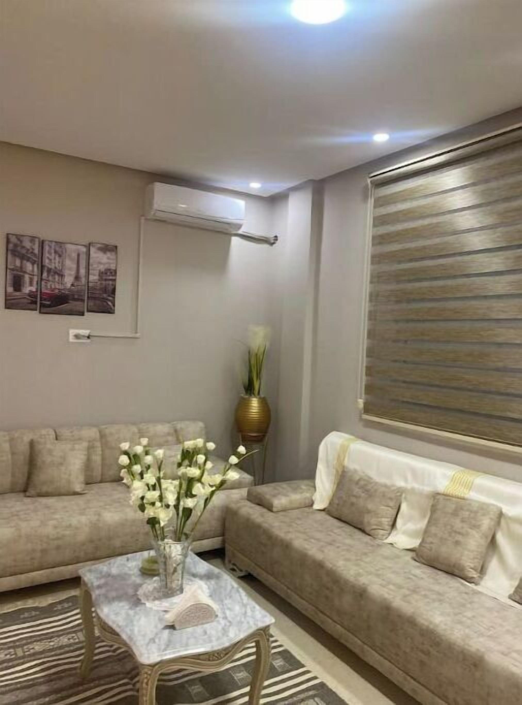 A living room with a beige sofa, a marble coffee table with a vase of flowers, a gold vase, and a large window with blinds.