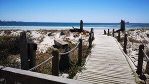 A wooden boardwalk leading to a beach with clear skies and distant ships. - Family Cape Town Beachfront Apartment (Cape Town)
