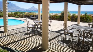 A covered outdoor seating area with a pool, chairs, and tables, overlooking a beach and mountains. - Family Cape Town Beachfront Apartment (Cape Town)
