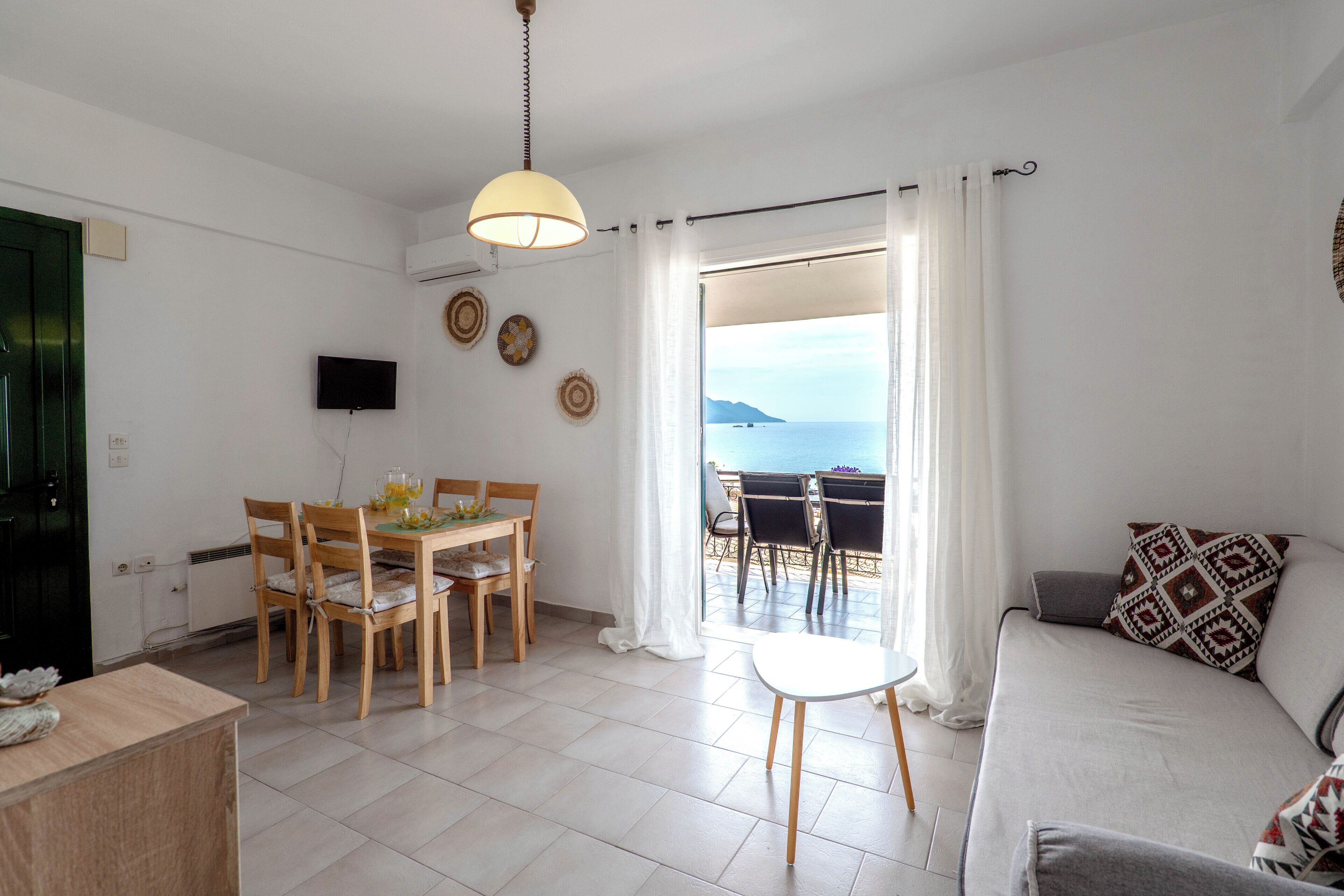 Una sala de estar con un sofá, una mesa de comedor con sillas, un televisor montado en la pared y vistas al mar a través de un ventanal grande.