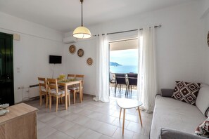 A living room with a sofa, a dining table with chairs, a TV mounted on the wall, and a view of the sea through a large window. - Corfu Glyfada Menigos 150 (Corfu)