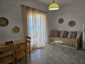 Ett rum med ett matbord, stolar, en soffa, ett litet runt bord, en gul hänglampa och ett fönster med tunna, genomskinliga gardiner.