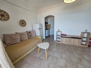 A living room with a sofa, a small white table, a kitchen in the background, and a wooden cabinet with shelves. - Corfu Glyfada Menigos 150 (Corfu)