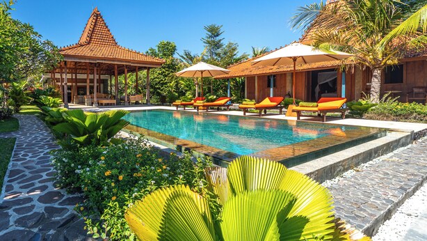Un espace piscine agrémenté de chaises longues et de parasols, entouré de plantes tropicales et d’un bâtiment de style traditionnel.